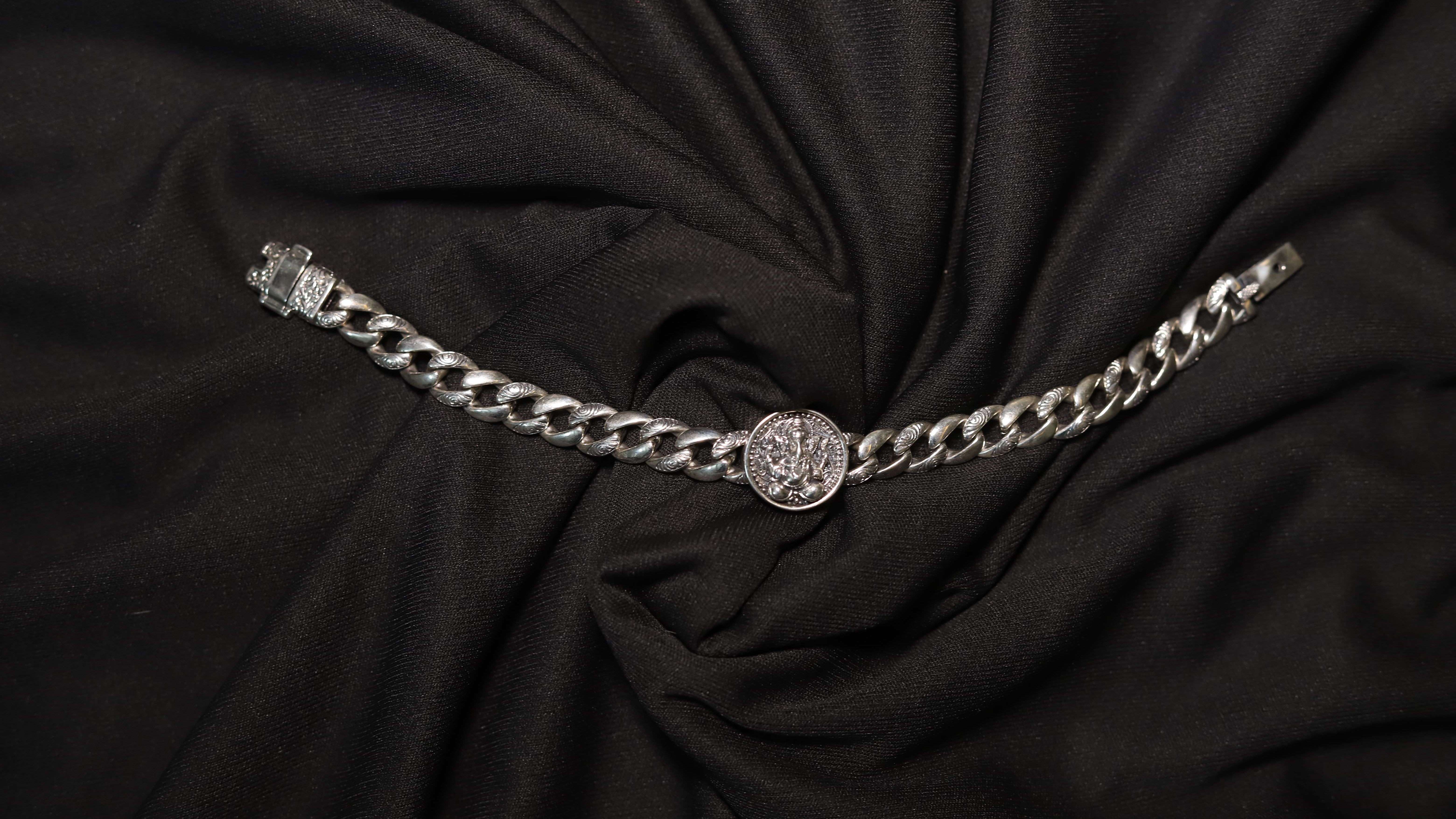 A silver-toned chain belt with a central round medallion and a clasp on the left, displayed on a draped black fabric background.